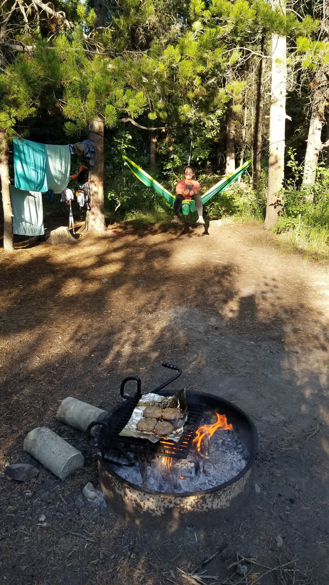 Burgers cooking on foil over a campfire with a green hammock strung between trees in the background