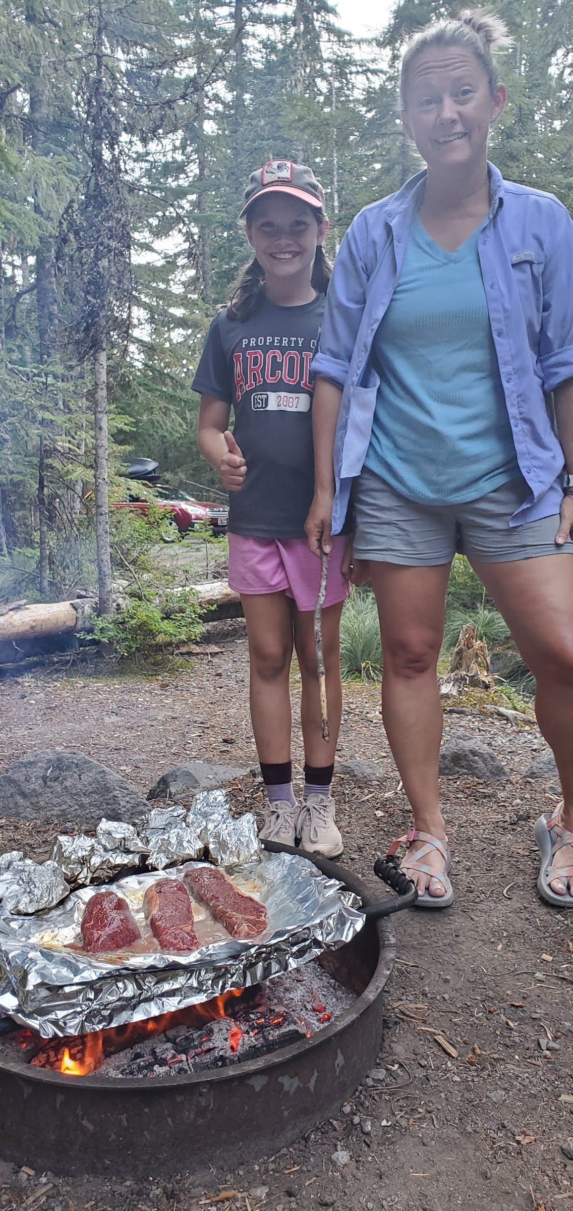 Woman and girl smiling behind steaks cooking on foil over a campfire