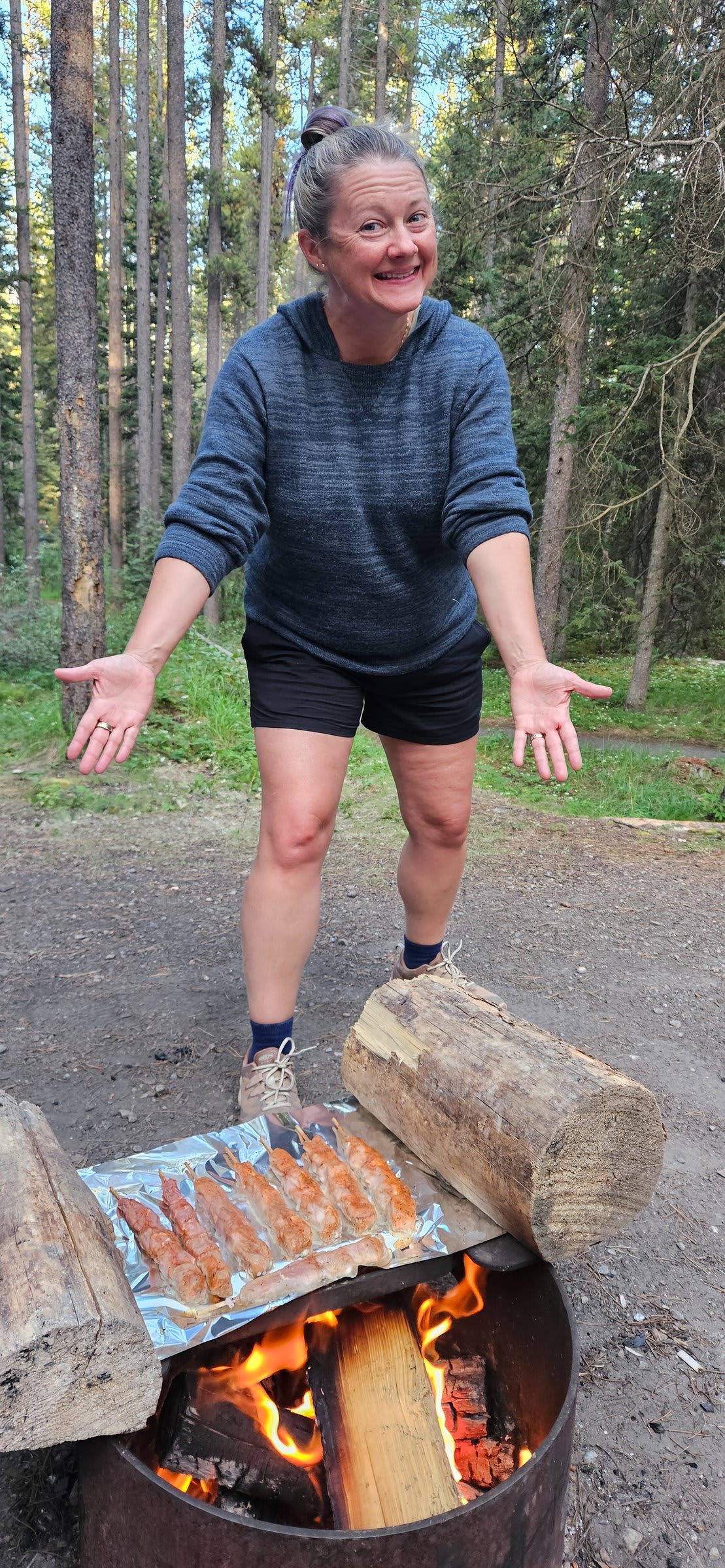 Woman smiling and gesturing toward meat cooking on foil over a campfire