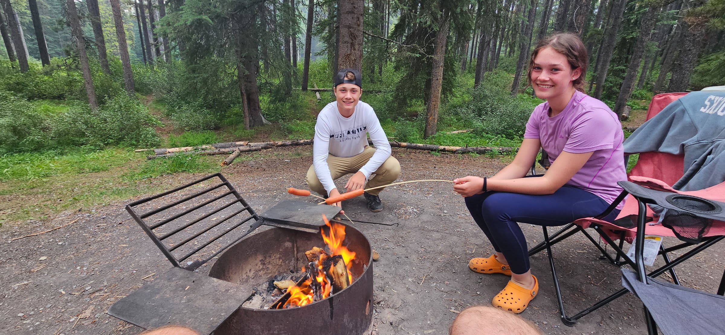 Two kids roasting hot dogs over a campfire and smiling at the camera