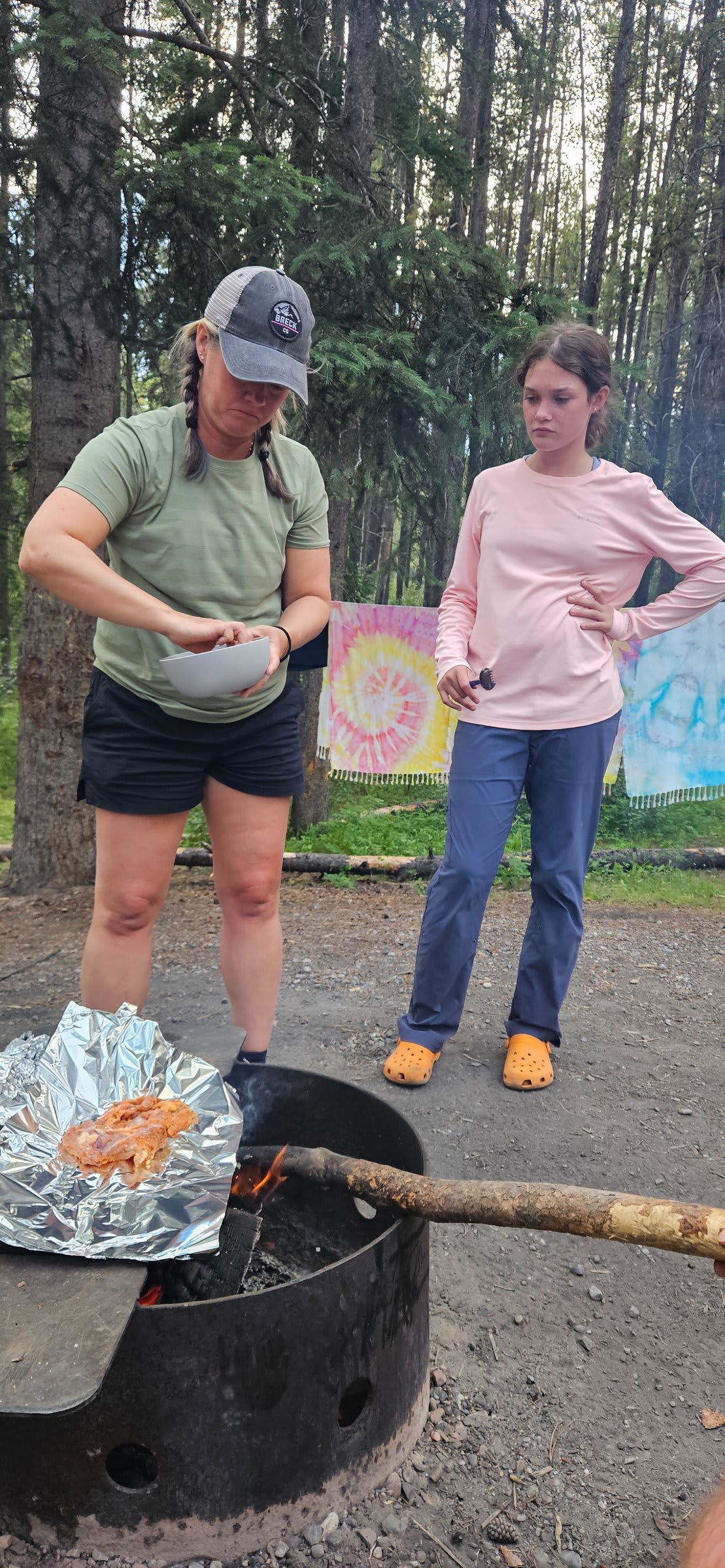 Two people standing by a fire ring watching marinated meat cook on foil