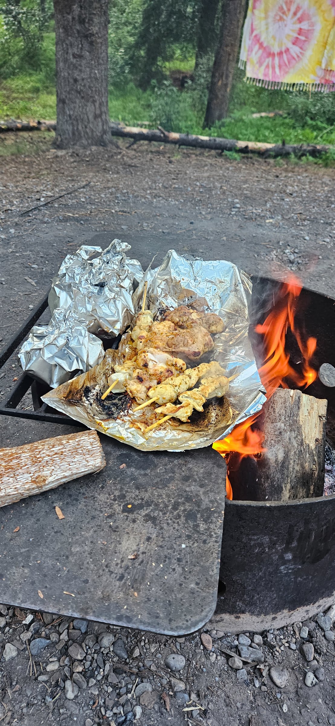 Seasoned chicken skewers and foil packets cooking on a grate over an open campfire