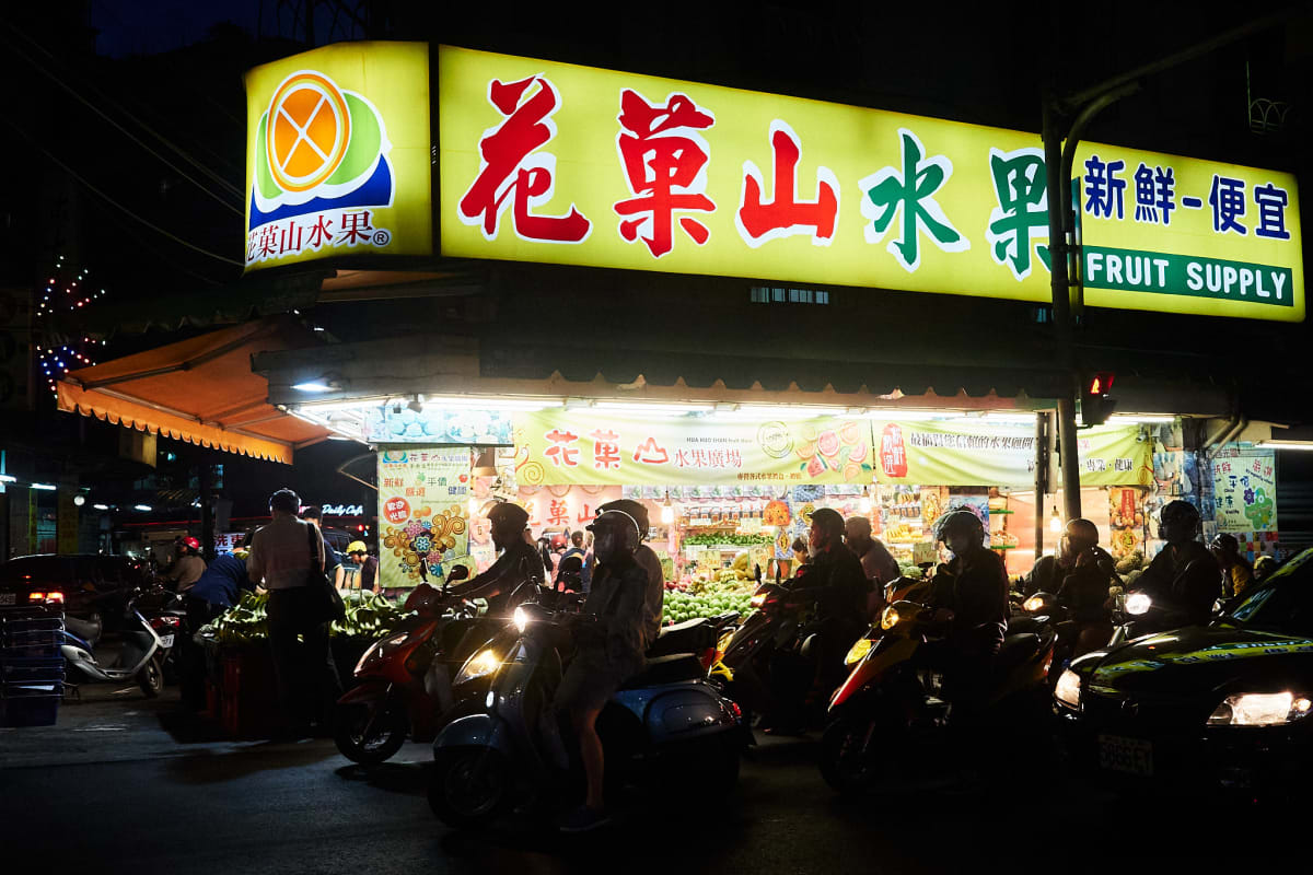 A fruit market with a row of scooters in front of it.