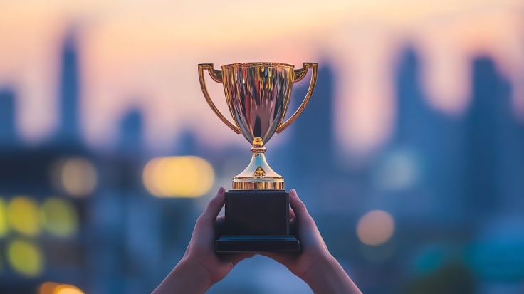 A person's hands hold up a gold trophy cup against a blurry cityscape background.