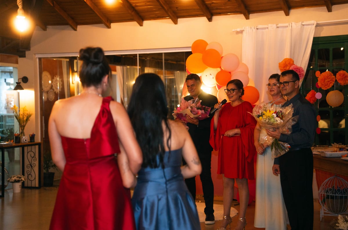 Convidados reunidos em uma festa no salão, com balões laranja e buquês