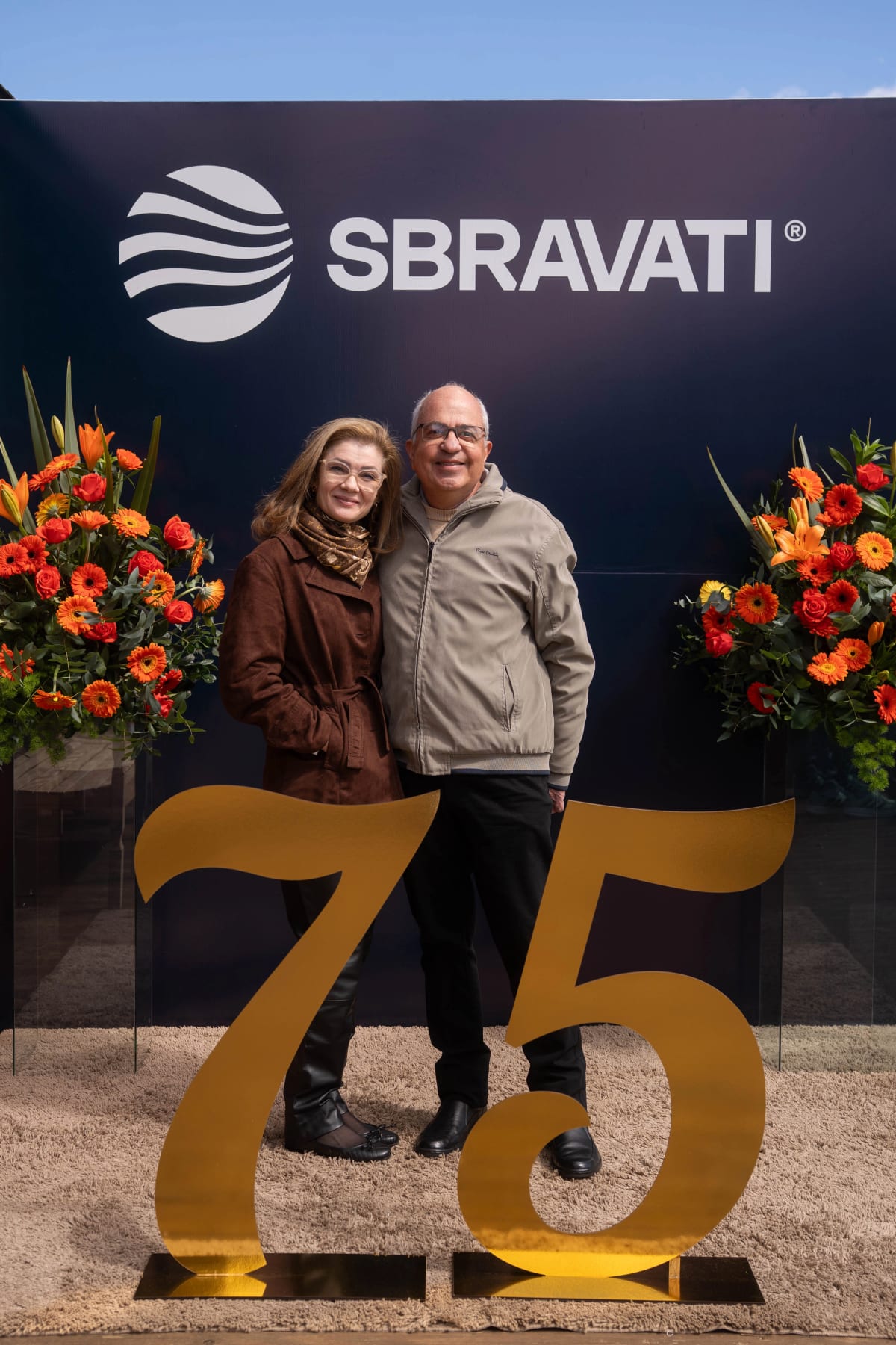 Casal de convidados abraçado diante do painel do evento de 75 anos