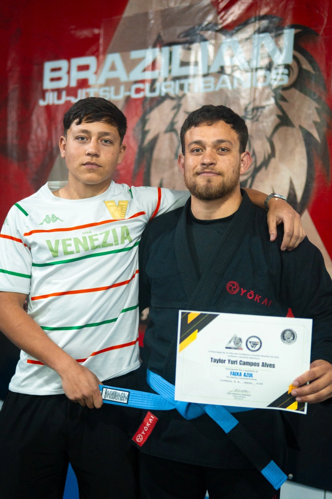 Dois alunos com o diploma de graduação de faixa azul diante do banner da academia