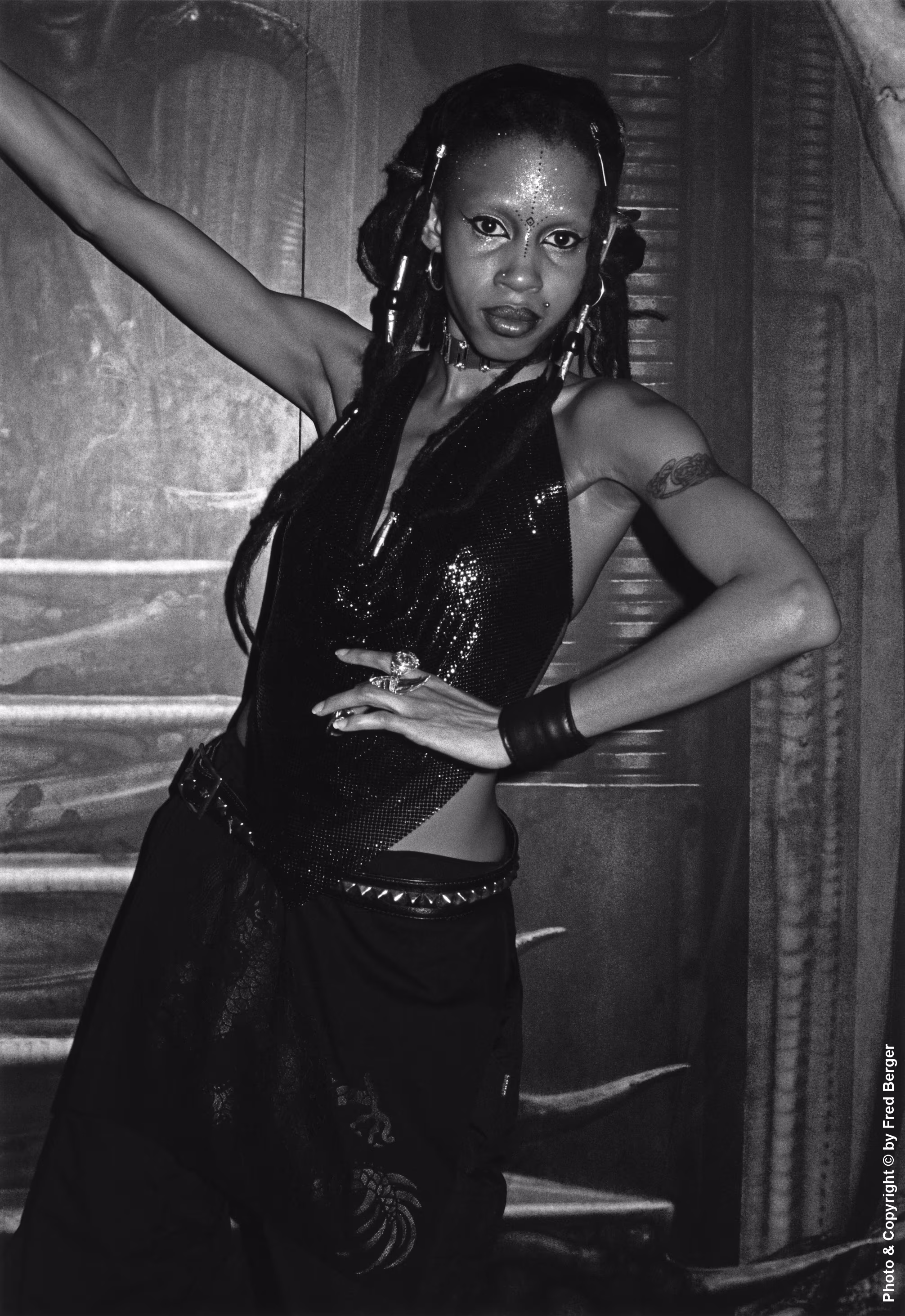 Keywords: Zen Warp, Zenwarp, Limelight nightclub, H.R. Giger Room, H.R. Giger, goth, gothic, cyber-goth, go-go girl, black goth, New York nightlife. This vertical Black & White photo depicts an attractive Black cyber-goth go-go girl. She&rsquo;s visible from the lower thighs up and is centered in the camera frame, which she fills completely from top to bottom. She&rsquo;s wearing a shiny halter top, cargo pants, metal studded leather belt, leather wristband, and leather choker - all in black. She has two ornate silver rings on her left hand. Her long black dreadlocks have four silver tubular clasps, two of which are on each side of her head. Her upper left arm has a tribal-style tattoo. Her body is tilted towards her left, with her right arm raised and her left arm bent towards her torso, as she looks at the camera. Her arms are slender with well-defined muscle tone. Her face is speckled with glitter, she has black eyeliner, silver eyeshadow, and her lips have a dark outline. Behind her is a mural by surrealist artist H.R. Giger depicting a science fiction style architectural structure. The photo is illuminated by a camera flash.
