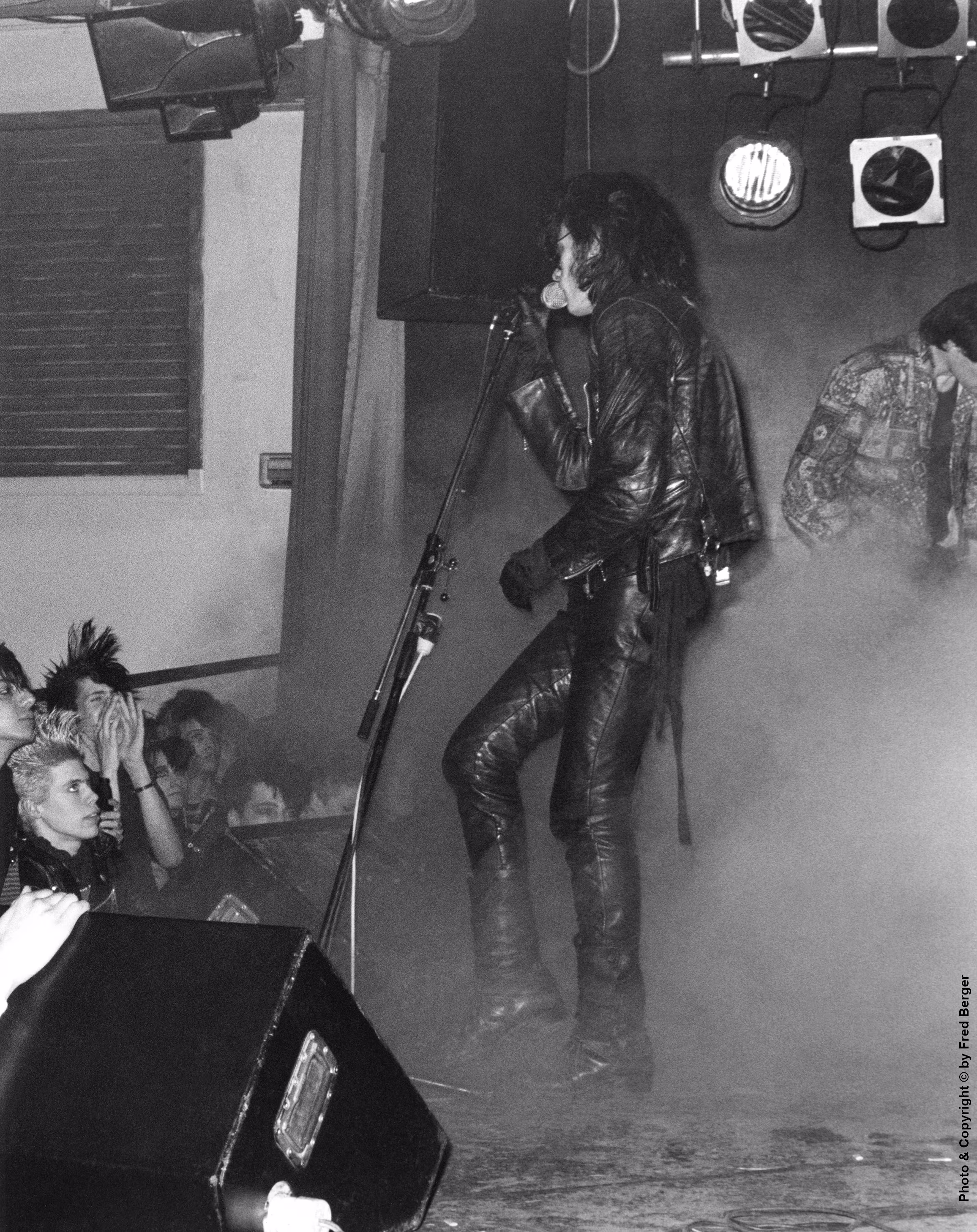 This vertical Black & White photo shows Andrew Eldritch of The Sisters of Mercy singing on stage at Danceteria on April 13, 1984. He wears a black leather jacket, leather pants, gloves, and boots, and holds the microphone to his mouth while standing in thick stage fog beside a microphone stand. A small group of punk fans, one with a mohawk and one with short blonde hair, look up at him from the lower left edge of the stage. Stage lights hang at the top right, and part of another performer is visible at the right edge. A credit reading Photo & Copyright &copy; by Fred Berger runs vertically along the lower right edge.