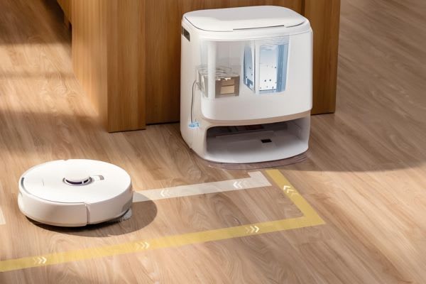 A robot vacuum docks by a charging station on wooden flooring, illustrating tips on how to hide robot vacuum devices in your home.