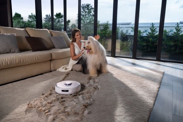 A woman pets her fluffy dog while a robot vacuum cleans up pet hair on the carpet, showcasing effective robot vacuum hacks.
