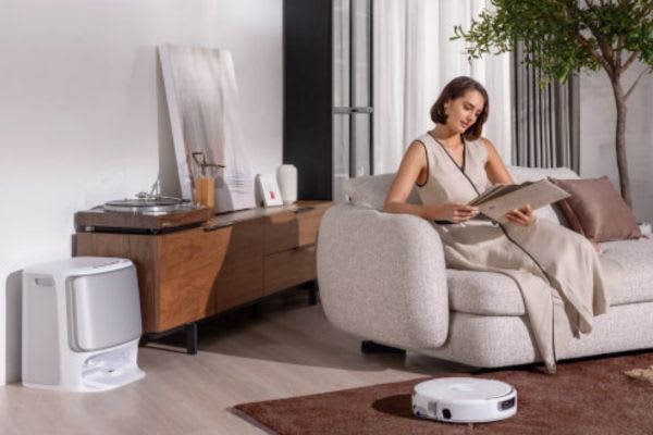 A woman sits comfortably on a sofa, enjoying a book while a robot vacuum operates on a stylish hardwood floor.