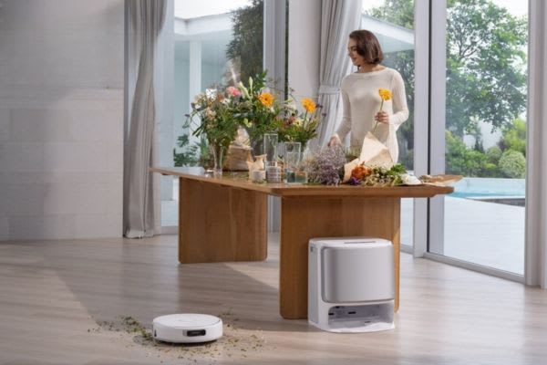 A woman stands by a wooden table with flowers, while a robot vacuum efficiently cleans the floor, showcasing its 5GHz capability.