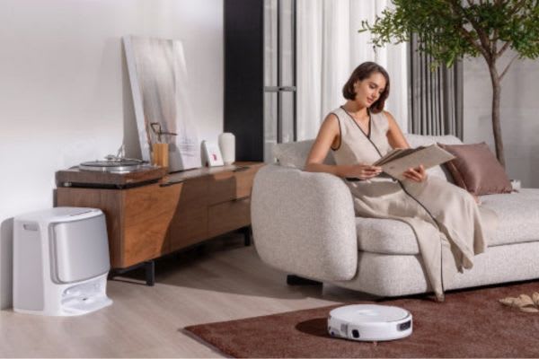 A woman reads calmly on a sofa while a robot vacuum moves on the floor, prompting the question: can robot vacuums go over threshold?