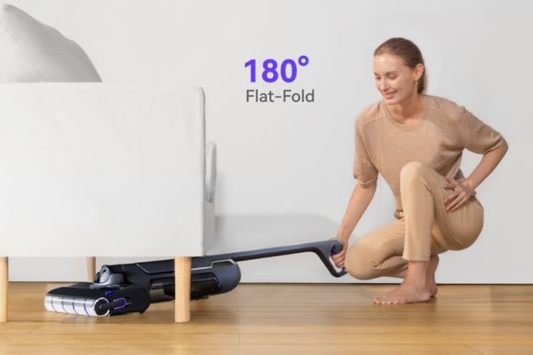 A woman kneels to clean under a couch, demonstrating how to get hair out of vacuum roller with a flat-fold vacuum design.