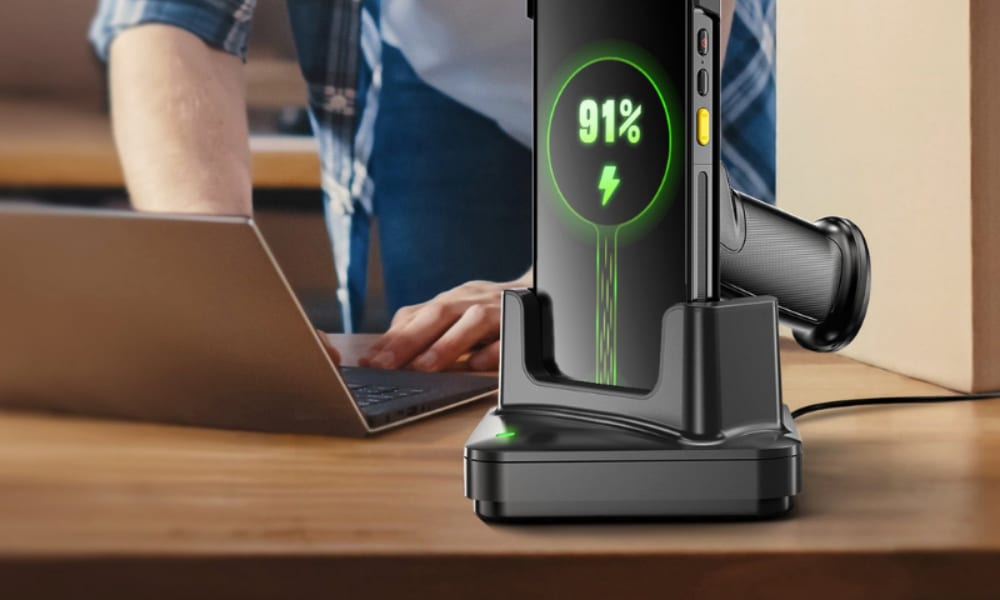 A warehouse scanner is docked and charging, showing 91% battery while a person works on a laptop nearby.