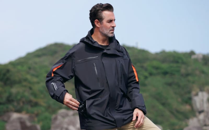 A man stands outdoors wearing a black jacket with orange accents, illustrating the differences between a coat vs jacket.