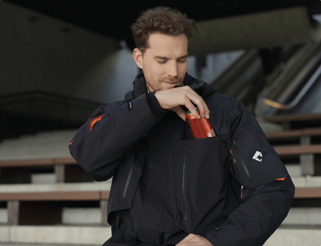 A person wearing a hardshell jacket opens a pocket to retrieve a drink, showcasing hardshell vs rain jacket features.