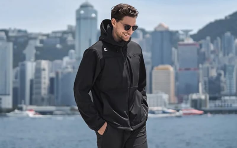 A man wearing a stylish black jacket stands near water, showcasing the debate of softshell vs windbreaker.