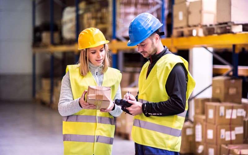 Two warehouse workers, one holding a package, discuss barcode scanner integration while wearing safety gear.