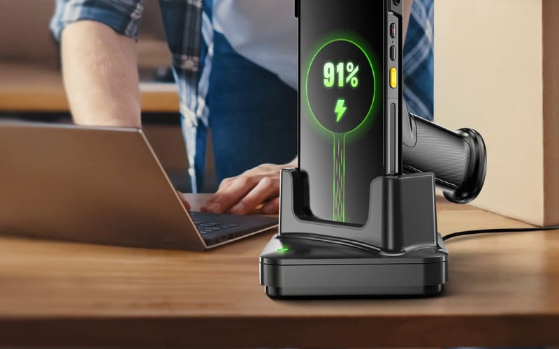 A close-up of a pistol grip scanner charging on a dock near a person working on a laptop, showcasing its sleek design.