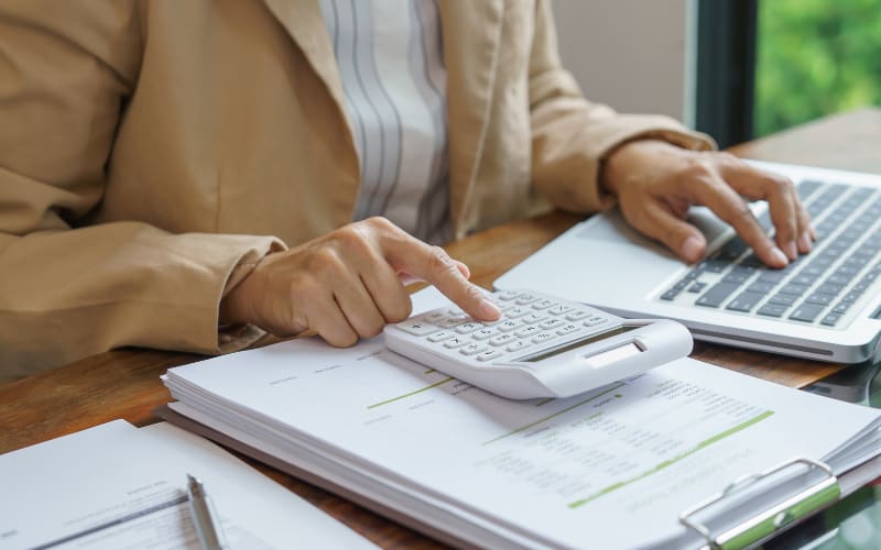 A person calculating with a calculator while working on financial documents, alongside a pistol grip scanner and a laptop.