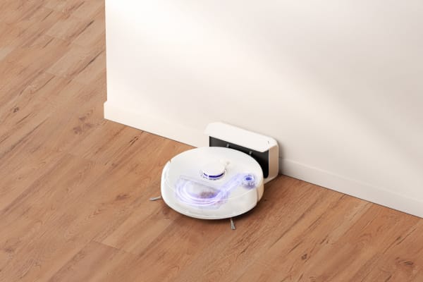 A robot vacuum with docking station positioned against a wall on a wooden floor, illuminated gently.