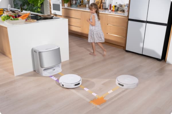 A robot vacuum with docking station is cleaning the floor in a bright kitchen while a child plays nearby.
