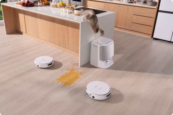 Two robot vacuums on a floor near a spilled drink, while a cat leaps over a kitchen counter. Can robot vacuums go over threshold?