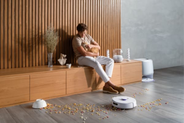 A person sits on a wooden bench with a dog, surrounded by scattered pet food, while a robot vacuum cleans nearby, raising the question: Can robot vacuums be used for pet hair removal?