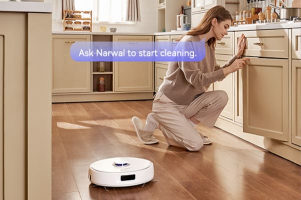 A woman in a kitchen uses a robot vacuum with remote control while kneeling on the floor.