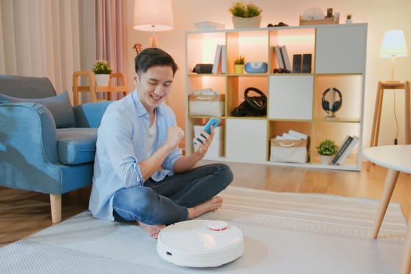A smiling man uses his phone while a robot vacuum cleans the floor, questioning: Do robot vacuums really clean your floors effectively?