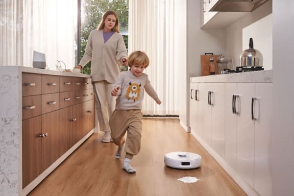 A mother and child playfully interact in a kitchen while a robot vacuum performs spot cleaning on the floor.