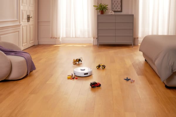 A robot vacuum on a hardwood floor surrounded by toy cars, demonstrating how to use your robot vacuum for daily cleaning routines.