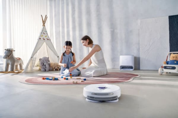 A mother and daughter play with blocks on a rug while a robot vacuum cleans nearby, showcasing how to use your robot vacuum for daily cleaning routines.