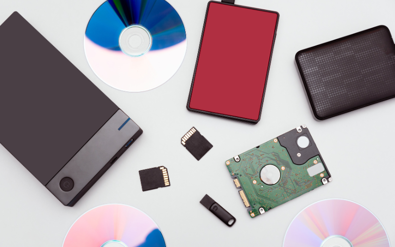 Various storage devices including hard drives, flash drives, discs, and memory cards arranged on a light background.
