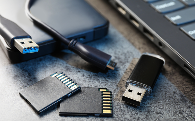 An assortment of storage devices including USB drives, SD cards, and a portable hard drive beside a laptop.