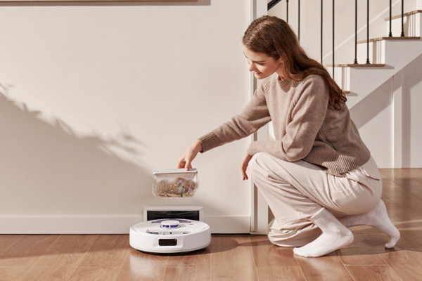 A woman kneels beside the thinnest robot vacuum, emptying its dust bin into a container, showcasing modern home cleaning.