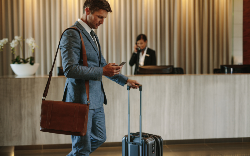 A businessman with a suitcase and a messenger bag checks his phone at a hotel reception, discussing removable battery options.