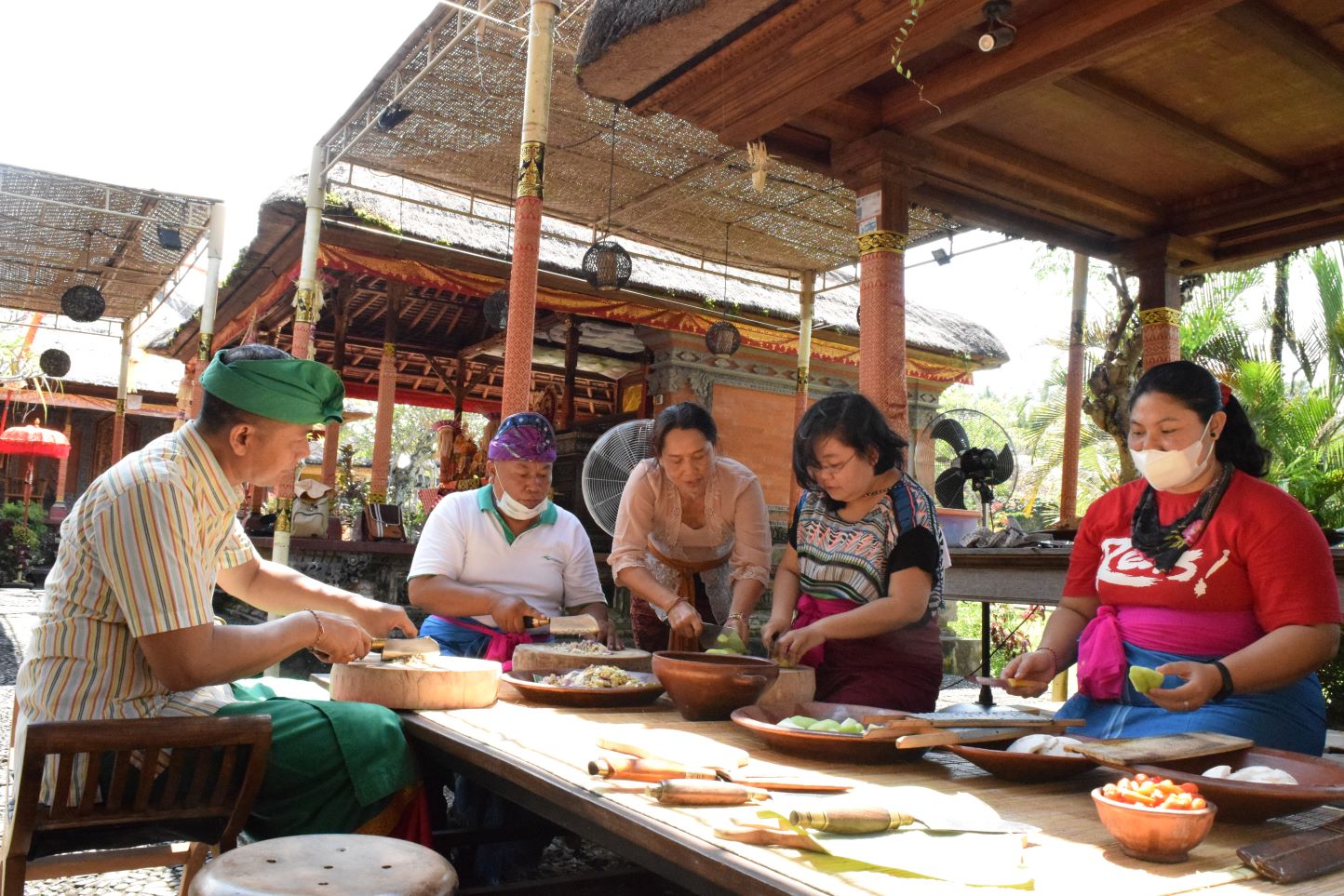 As the gathering begins, a Balinese community comes together under a traditional pavilion to prepare food and share daily activities, reflecting local culture and togetherness.