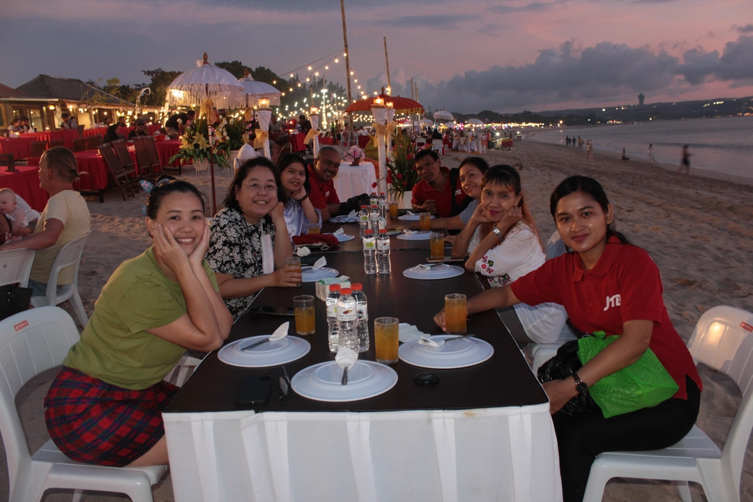 As dusk settles, guests enjoy beachfront dining in Bali with a shared table set on the sand, warm lights overhead, and the ocean creating a relaxed evening atmosphere.