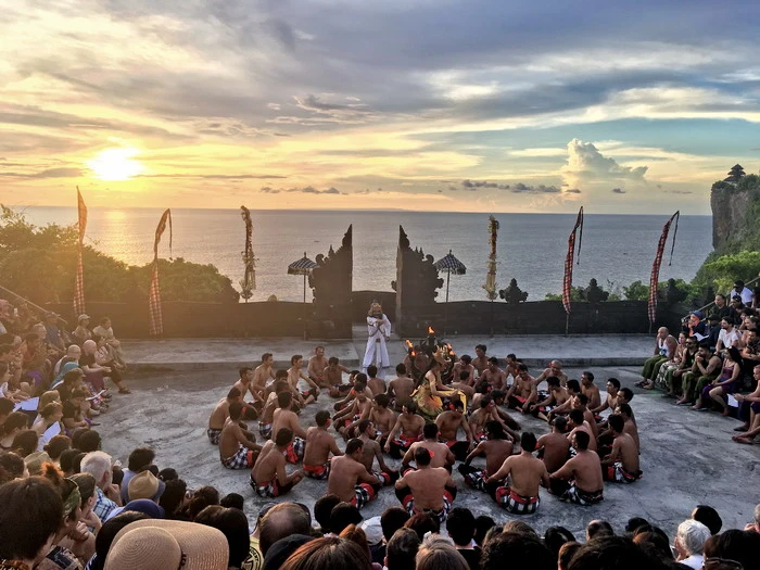As evening approaches, a traditional Kecak dance performance takes place at an open-air temple in Bali, with dancers encircling a fire against a dramatic sunset backdrop overlooking the ocean.