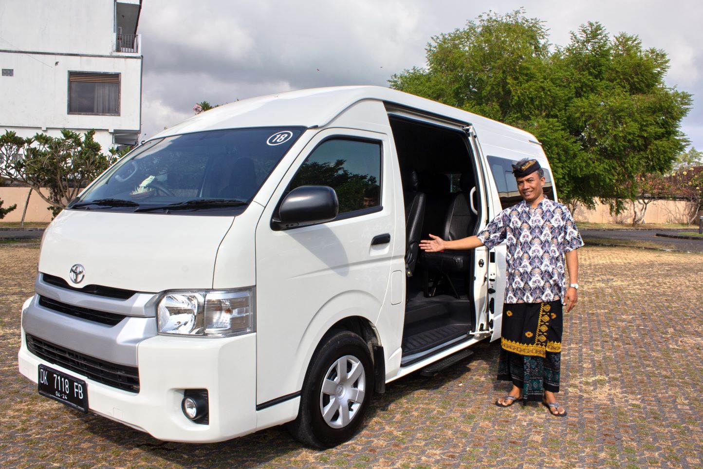 A JTB transport driver in Balinese attire stands beside a white Toyota HiAce with the sliding door open, welcoming guests for a private transfer service in Bali.
