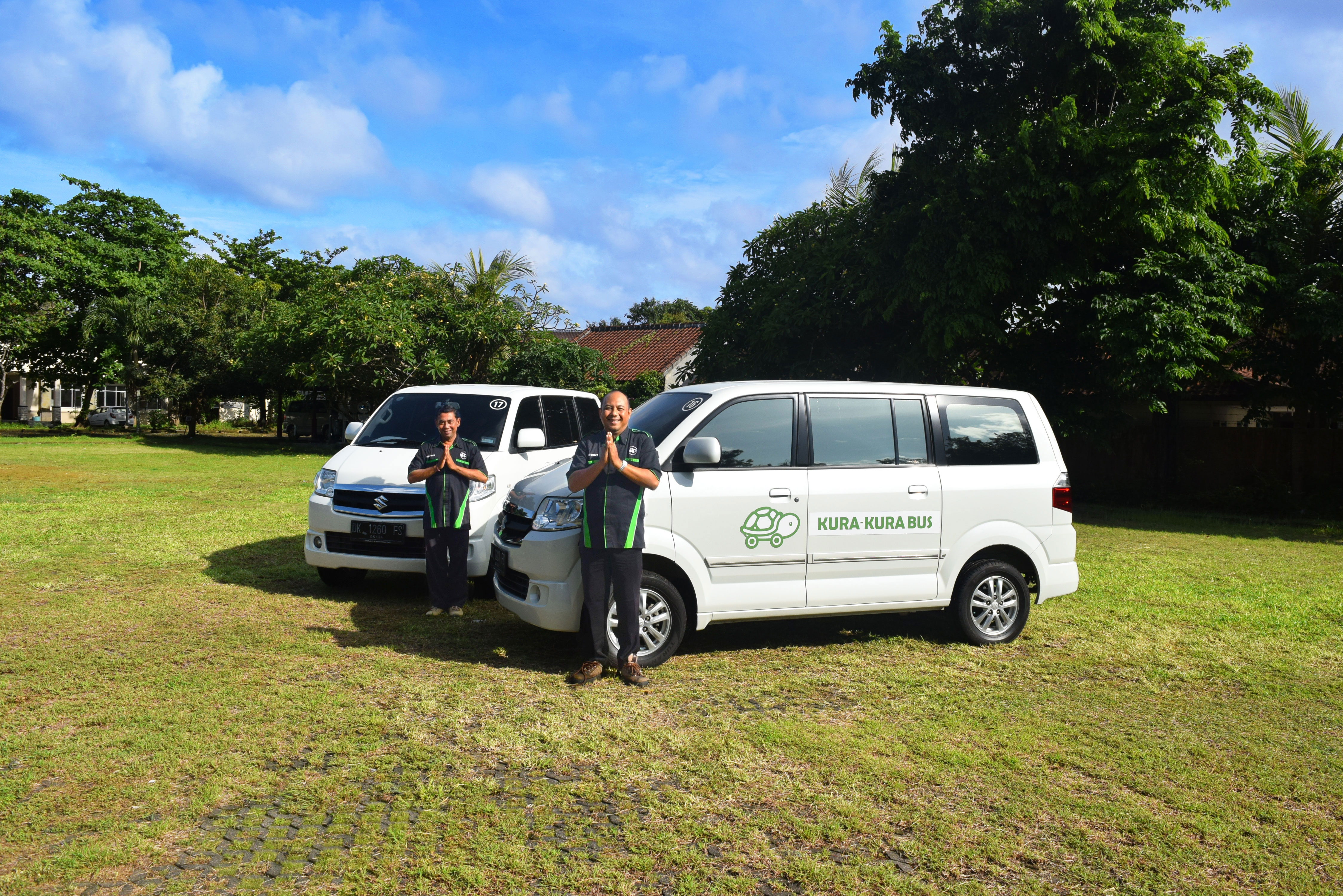 Meanwhile, two Kura-Kura Bus staff stand in front of white vans on a sunny grassy field.