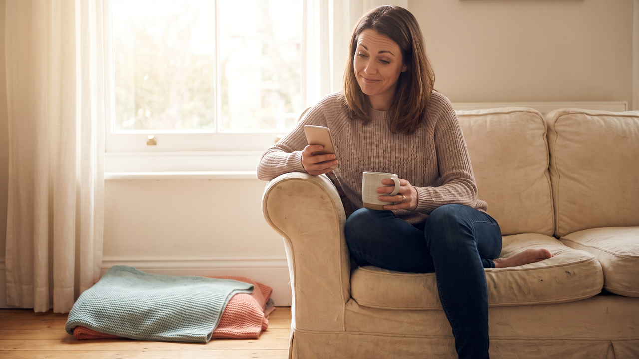 A physician sitting on a sofa arm in morning light, reading a bonus pay stub on her phone with a coffee mug in hand