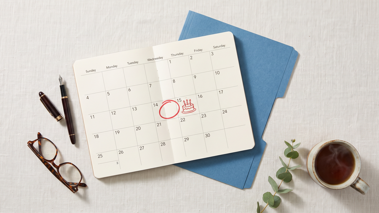 Overhead view of a calendar with a birthday circled next to a health insurance enrollment folder, pen, and reading glasses