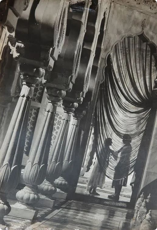 Silhouettes of a woman and a child holding hands in an ornate corridor with decorative pillars and draped fabric.