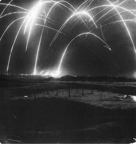 Black and white image of bright arcs of light in the night sky, resembling artillery fire or fireworks, with a dark landscape below.