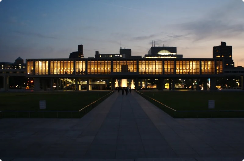 A modern building with large illuminated windows, set against a twilight sky, with a pathway leading towards it and people walking in the foreground.