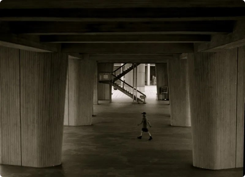 A black and white photograph of a person walking beneath large concrete pillars, with a staircase visible in the background.